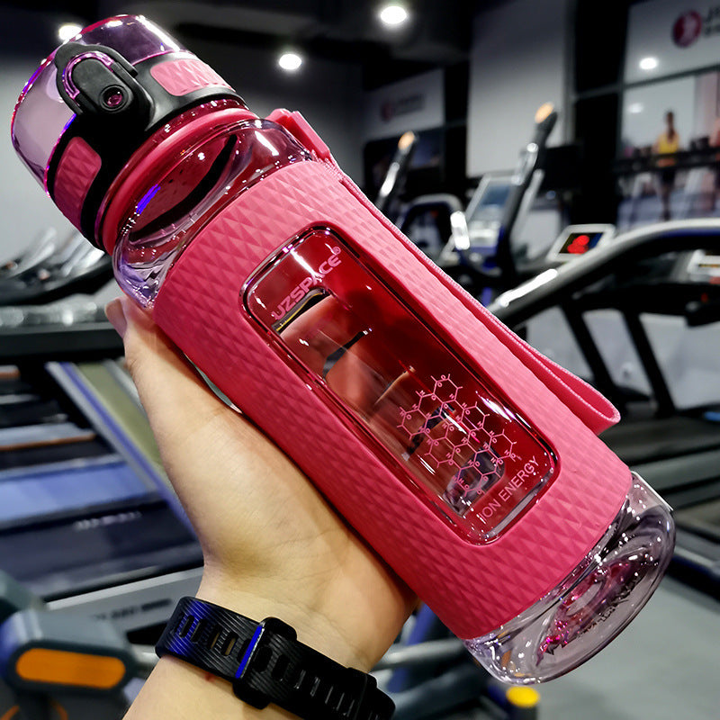 Pink water bottle with a clear section held in a hand, gym equipment in the background