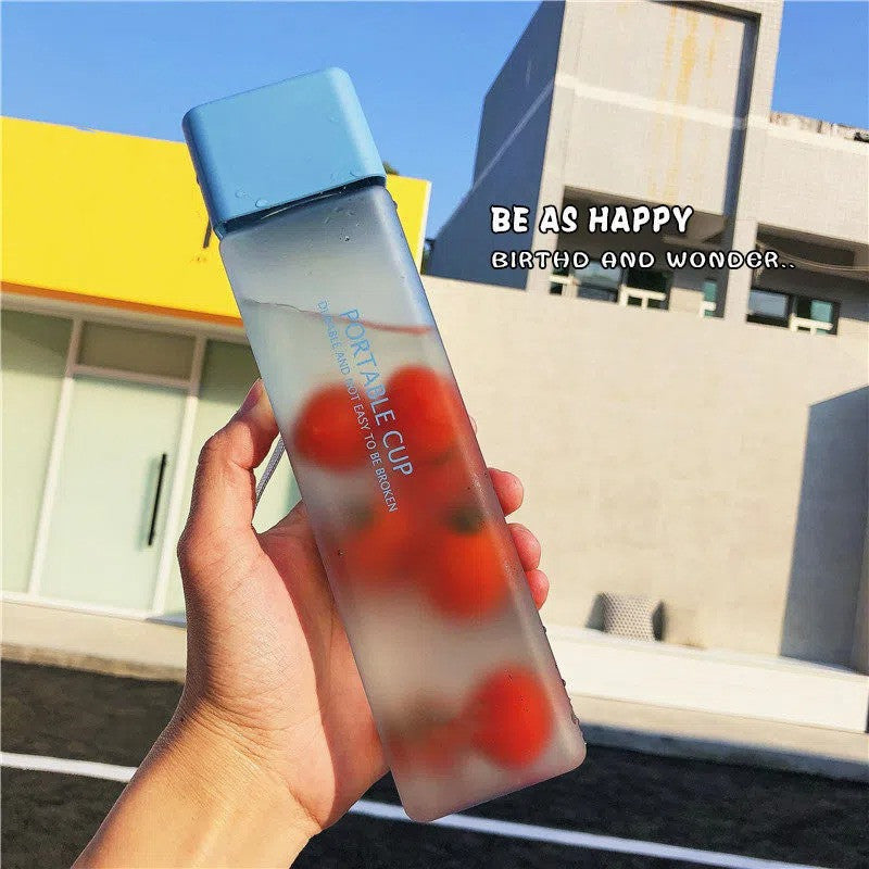 Hand holding a transparent cup with red fruit pieces and blue lid, with 'Be as Happy' text in the background.