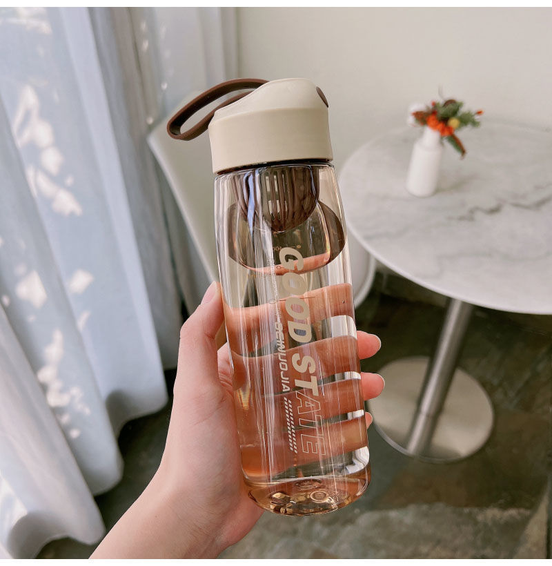 Clear water bottle with brown lid held by a hand, blurred indoor background