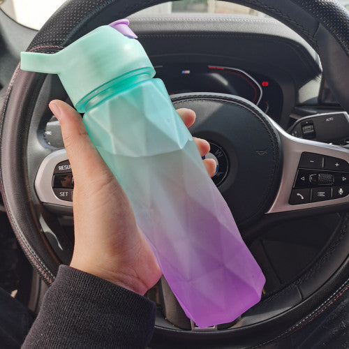 Hand holding a gradient green to purple water bottle in front of a car steering wheel.
