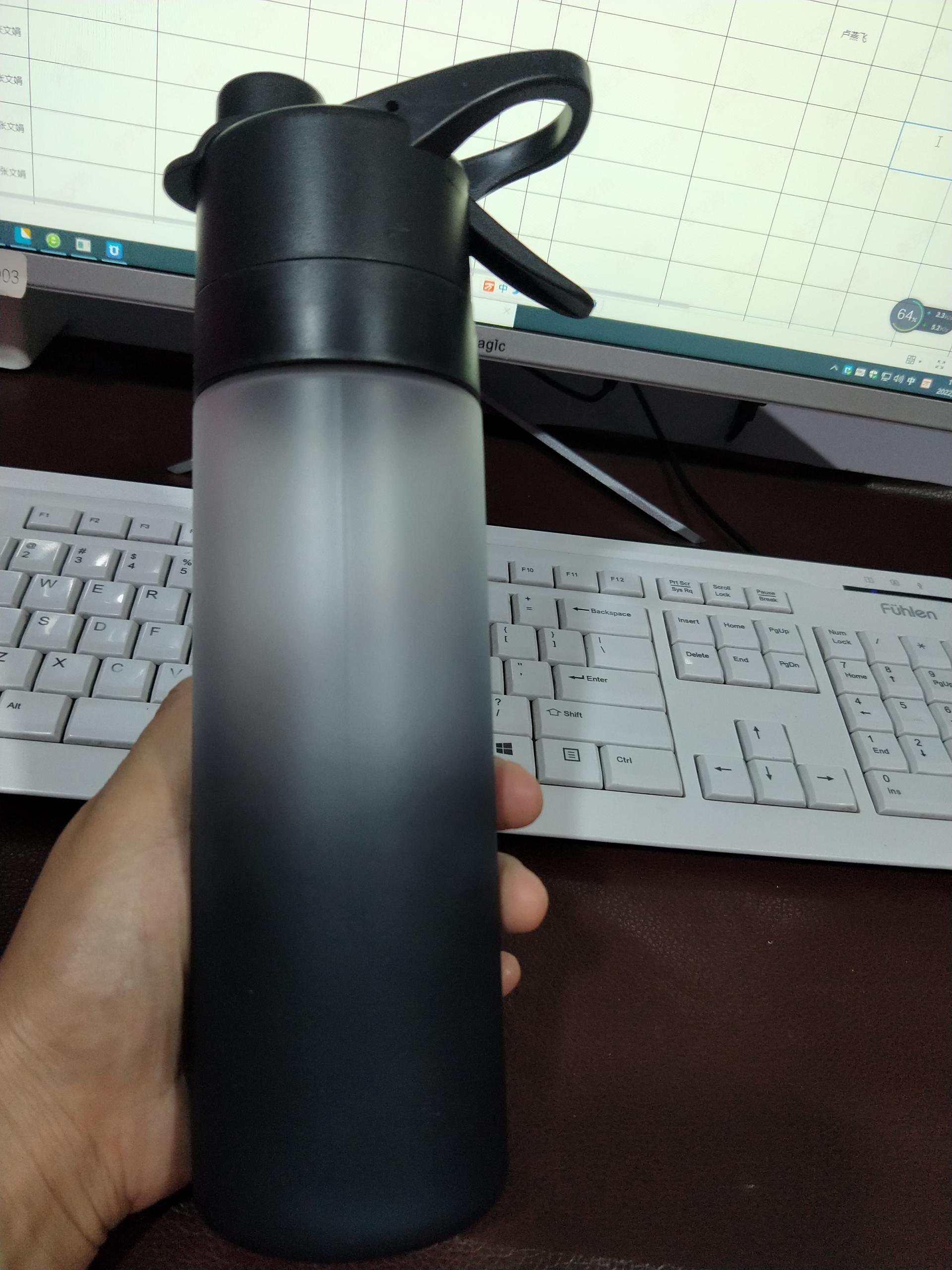 Black water bottle held in a hand with a keyboard and monitor in the background