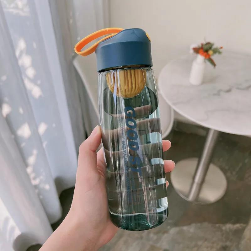 Clear water bottle with blue cap and orange handle held by a hand, blurred indoor background
