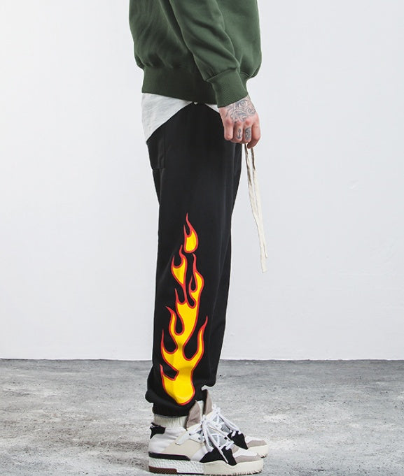 Person wearing black sweatpants with flame design, green hoodie, and white sneakers on a light gray background.