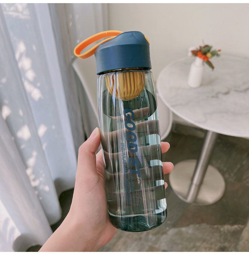 Clear water bottle with blue cap and orange handle held by a hand indoors.
