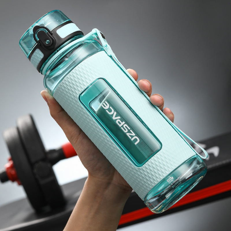 Hand holding a light blue water bottle with 'UZSPACE' branding, gym equipment in the background